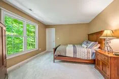 Bedroom interior with a queen-sized bed, wooden furniture, and a large window with a view of green foliage.