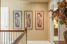 Hallway in a home featuring three framed artworks on the wall, a wooden banister on the left, and a floral arrangement in the foreground on the right.
