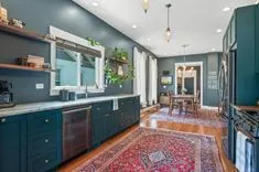 Elegant interior of a spacious kitchen with dark green cabinetry, stainless steel appliances, Persian rug on the hardwood floor, leading to a well-lit dining area.