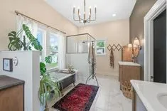 Elegant bathroom interior with a freestanding bathtub, separate shower area, double vanity sinks, and a chandelier, accented by indoor plants and a red area rug.