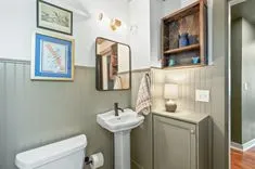 Small bathroom with pale green wainscoting, white fixtures, decorative towel, framed pictures, and a shower door glimpsed on the right.