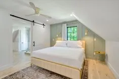 Bright and airy attic bedroom with a queen-sized bed, sliding barn door leading to an en suite bathroom, hardwood floors, and a large window with a view of greenery.