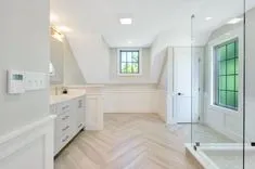 A bright, contemporary bathroom with white cabinetry, herringbone patterned wood floor, dormer window, and a large mirror reflecting a green window pane.