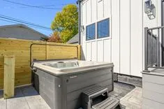 Outdoor residential hot tub with a closed cover on a wooden deck next to a wooden privacy fence, with a white exterior wall and windows in the background, on a sunny day.