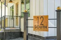 A wooden house number sign reading "12 SOUTH" and an Instagram handle "@thathouseon12south" mounted on a dark metal post at the entrance of a home with a white door and sidelights in the background.