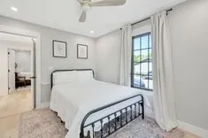 Bright modern bedroom with a metal frame bed, white bedding, light gray walls, two framed botanical prints, a ceiling fan, and curtains drawn back to reveal a sunny window.