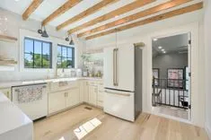 Bright modern kitchen with wooden beams, white cabinetry, stainless steel appliances, and an open doorway leading to an adjacent room.