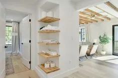 Modern home interior with wooden floating shelves storing towels and toiletry items, a cozy living room with armchairs in the background, and exposed wooden beams on the ceiling.
