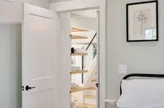 Interior view of a modern bedroom with an open door showing a glimpse of a staircase and shelving unit, with a framed picture on the wall.