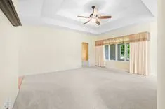 Spacious empty room with beige walls and carpet flooring, a ceiling fan, and large windows covered by light curtains.