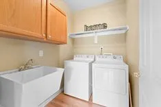 A laundry room with a washing machine, dryer, utility sink, wooden cabinets, and a decorative sign that reads 'home'.