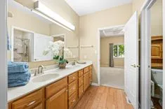 A well-lit, spacious bathroom with a large vanity, two sinks, wooden cabinets, a mirror reflecting a glass-door shower, and an open door leading to an adjacent room.
