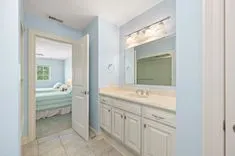 Bright bathroom with blue walls, white cabinetry, and a large mirror over a sink, with a view into an adjoining bedroom.