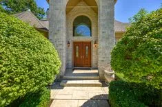 Elegant house entrance with stone columns, arched doorway, and wooden double doors flanked by trimmed green bushes under a clear blue sky.