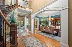 Elegant interior view of a house featuring a curved wooden staircase with ornate metal balusters, archway to a dining room with a chandelier and a set dining table, and through another arch a glimpse of a living room area.