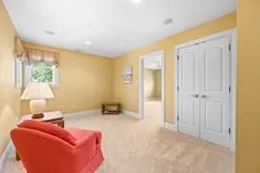 Brightly lit room with yellow walls, a coral chair, white doors, and a beige carpet, with an open doorway leading to another room.
