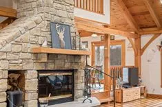 Interior of a cozy cabin living room with a large stone fireplace, wooden beams, and glass doors leading outside.