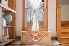 Cozy wooden interior with a plush fox toy sitting on a cushion inside a handmade wooden crate, beside a sunny window draped with sheer curtains, with a globe on a shelf and wooden stairs to the right.