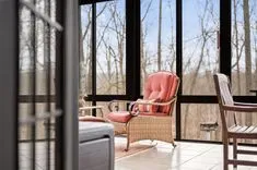 A cozy sunroom with a wicker armchair with red cushions overlooking a forest area, sunlight streaming through large windows, and a wooden deck chair in the background.