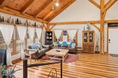 Spacious living room with wooden ceiling beams, hardwood floors, large windows with sheer curtains, and rustic furniture including a couch, chairs, and wooden coffee table.