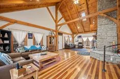 Spacious living room with high wooden ceilings, exposed beams, hardwood floors, a stone fireplace, and comfortable leather furniture.