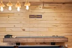 Interior of a game room with a shuffleboard table in focus, pine wood wall in the background, and a sign that reads "Play Shuffleboard Here" under warm hanging lights.