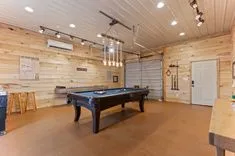 Spacious game room with a pool table, wooden walls, modern hanging light fixtures, and an industrial-style roll-up door.