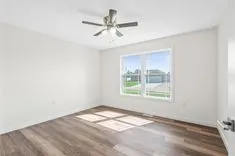 Empty room with wood flooring, white walls, ceiling fan, and a window with a view of a suburban neighborhood.