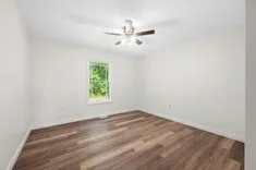 Empty room with white walls, wooden flooring, a window with a view of trees, and a ceiling fan with lights.