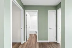 Interior view of a clean, brightly lit hallway with wooden flooring, sage green walls, and multiple white doors, one of which is open to reveal a bathroom with a white tub.