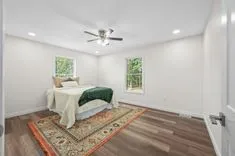 Bright minimalist bedroom with a bed, ceiling fan, recessed lighting, two windows, and a traditional area rug on a hardwood floor.