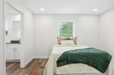 Bright modern bedroom with white walls, a bed with cream bedding and a green knit blanket, wooden flooring, a window with a view of greenery, and an attached kitchenette.