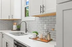 Bright modern kitchen with white cabinets, subway tile backsplash, and stainless steel sink under a window with greenery outside.