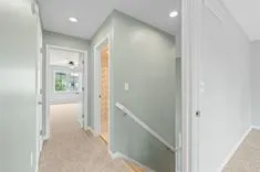 Bright hallway in a modern home with beige carpeting and white walls, featuring a railing and multiple doors leading to different rooms.