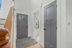 Bright interior hallway with wooden stair railing on the left, two gray doors, and a small window showing outdoor greenery.