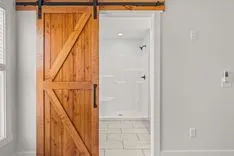 A sliding barn door crafted from warm-toned wood partially open to reveal a modern bathroom with white tiling.