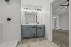Modern bathroom interior with dual sinks, large mirror with lighting, gray vanity, shower unit, and view into adjoining room with carpeted floor.