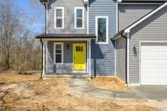 A new two-story house with gray siding and a bright yellow front door, featuring a white garage door and a concrete walkway in a barren front yard.