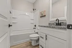 A modern bathroom interior with a white bathtub and shower combo, toilet, and vanity with granite countertop and framed mirror.