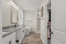 Modern bathroom interior with double vanity, granite countertop, large mirror, freestanding tub, decorative wall art, and a view into a walk-in closet.