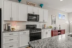 Modern kitchen interior with white cabinetry, stainless steel appliances, and granite countertops. Decorative accents include a 'COFFEE' sign and pendant lights.
