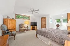 Bright bedroom with a large bed, ceiling fan, wooden furniture, and multiple windows letting in natural light.