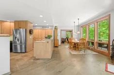 Modern kitchen interior with stainless steel appliances and wooden cabinets, adjacent to a dining area with a table set near large windows overlooking greenery.