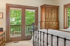 Cozy bedroom interior with iron bed frame, wooden furniture, balcony doors, and view of greenery outside.