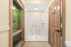 Interior of a modern bathroom with a large walk-in shower, wooden doors, and a window overlooking greenery.