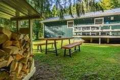 Wooden picnic table and pile of chopped firewood next to a green cabin with a porch in a forested area.