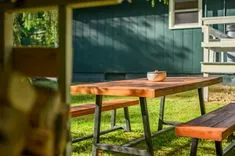 Wooden picnic table with bench in a sunny backyard with a bowl on the table and a green house in the background.