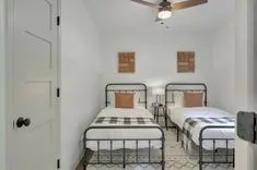 A neatly furnished bedroom with two twin beds with plaid bedding, a shared nightstand with a clock, framed artwork on the walls, and a ceiling fan above.