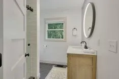 Modern bathroom interior with a wooden vanity, white sink, large round mirror, a window with greenery outside, white subway tiles in the shower, and a patterned rug on the floor.