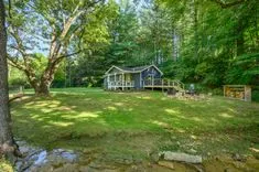 Charming blue cottage with a white porch and wooden deck surrounded by lush trees and a well-kept lawn with a swing set and fire pit area near a small brook.